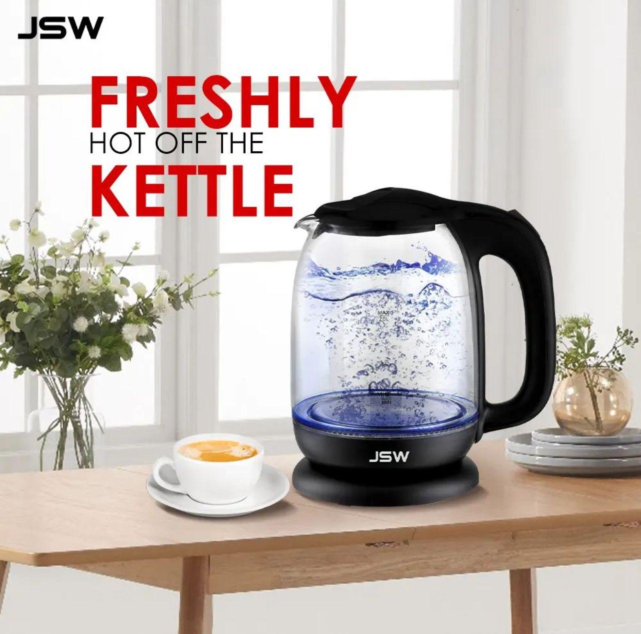 Glass electric kettle boiling on a wooden table next to a cup of coffee and white flowers.